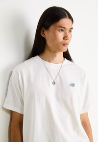Bílá bavlněná tričko s malým šedým logem "New Balance" na levé části hrudníku, doplněné stříbrným náhrdelníkem s přívěskem slunce.
