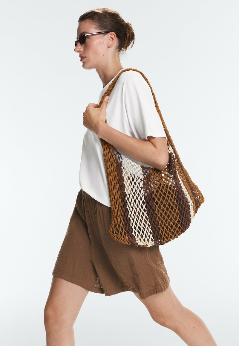 Femme portant des lunettes de soleil, chemise blanche, short marron, portant un grand sac en filet marron et crème sur l'épaule, marchant devant un fond uni.