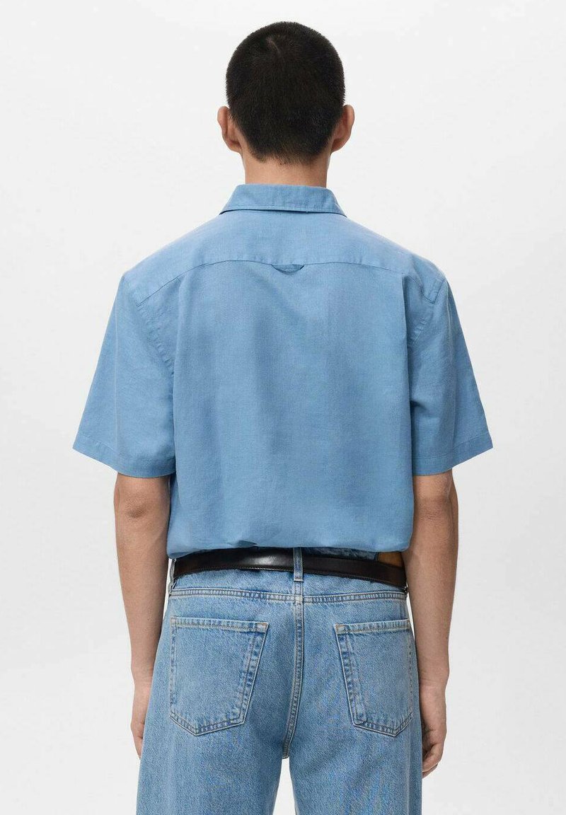 Leicht hellblaues Hemd mit kurzen Ärmeln aus Leinen, mit einem Kragen und einem rechteckigen Passe. Kombiniert mit hellblauen Jeans und einem schwarzen Gürtel.