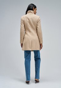 Mujer con cabello largo y negro que lleva un abrigo beige con botones, vaqueros azules y sandalias negras de tacón, de pie frente a un fondo gris liso.