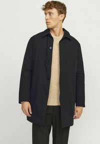 Abrigo negro de manga larga con diseño de botones en la parte delantera, cuello con muescas y dos botones. Lleva puesto un suéter beige claro. Textura de tela suave.