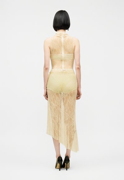 Γυναίκα με κοντά μαύρα μαλλιά, φορώντας διάφανο μπεζ δαντελένιο φόρεμα και μαύρα τακούνια, στέκεται με την πλάτη στραμμένη προς τα εμπρός σε απλό λευκό φόντο.