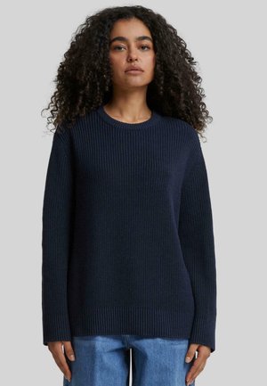 Navy knit sweater with a ribbed texture, round neck, long sleeves, and a relaxed fit, paired with light blue denim pants.