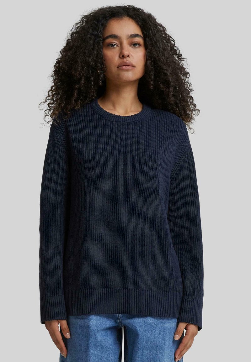 Pull en mailles bleu marine avec une texture côtelée, col rond, manches longues et une coupe décontractée, associé à un pantalon en denim bleu clair.