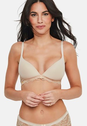 Femme aux longs cheveux foncés portant un soutien-gorge en dentelle beige et une culotte assortie en dentelle, debout avec les mains délicatement jointes à la taille.