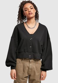 Ung kvinne med krøllete hår iført en kort svart knappet cardigan, tykk sølvkjede og løse beige bukser.