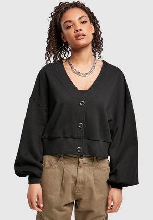 Ung kvinne med krøllete hår iført en kort svart knappet cardigan, tykk sølvkjede og løse beige bukser.