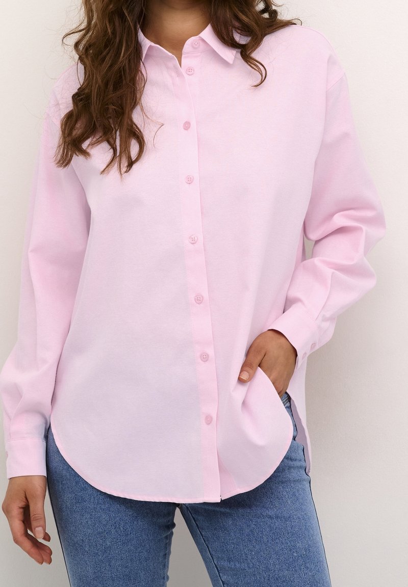 Femme portant une chemise rose clair à manches longues boutonnée, une main dans la poche, associée à un jean bleu sur un fond uni.