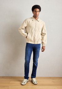 Beige Kapuzenjacke mit elastischen Bündchen, kombiniert mit dunkelblauen Jeans und grünlichen Turnschuhen, stehend auf einem Holzboden vor einer hellen Wand.