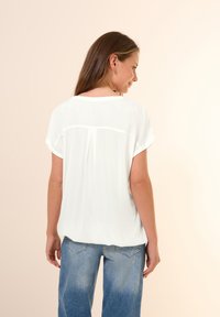 Blusa blanca de manga corta con una textura ligeramente translúcida y un plisado en la parte posterior. Combinada con jeans de corte relajado en azul claro.