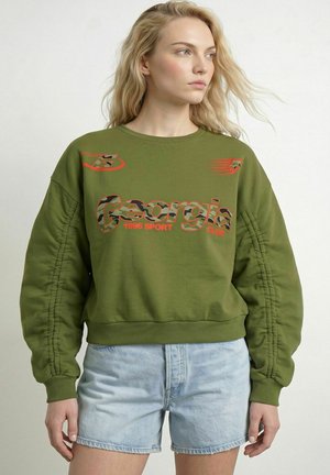 Blondynka w oliwkowej bluzie z napisem „Georgia 1996 Sport Club” i jasnoniebieskich szortach jeansowych na jednolitym tle.
