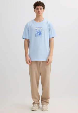 Joven de pie con camiseta azul claro que dice "Whitehall Rowing Club", pantalones beige con bolsillos parche y zapatillas blancas sobre fondo liso.