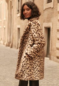 Kabát z falošnej kožušiny s leopardím vzorom v béžovej a hnedej farbe, vo voľnom strihu a oversized dizajne, umiestnený na dlažobnej ulici.