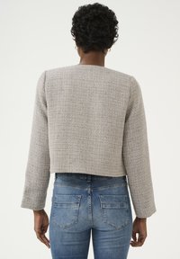 Szürke tweed cropped dzseki hosszú ujjakkal, texturált anyagból, négyzet alakú dekoltázzsal és látható varrással, kék farmernadrággal párosítva.