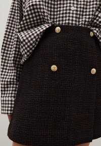 Camisa de cuadros marrón de botones y manga larga, combinada con una falda negra texturizada con botones dorados en el frente.