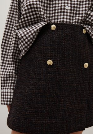 Braun kariertes Hemd mit langen Ärmeln, kombiniert mit einem strukturierten schwarzen Rock, der an der Vorderseite goldene Knöpfe hat.