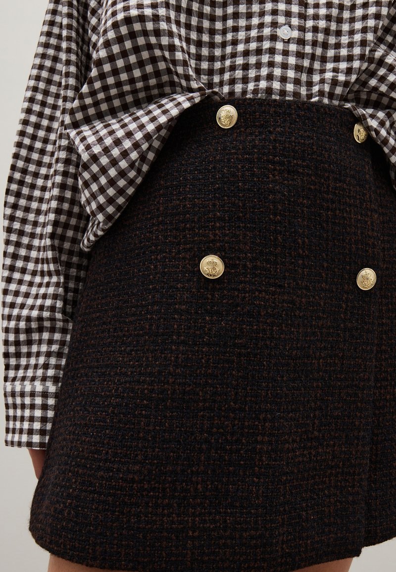 Camisa de cuadros marrón de botones y manga larga, combinada con una falda negra texturizada con botones dorados en el frente.