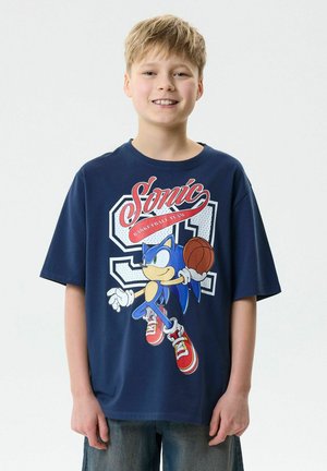 Garçon souriant, portant un t-shirt bleu marine Sonic the Hedgehog, tenant un ballon de basket, et un jean bleu, debout devant un fond clair uni.