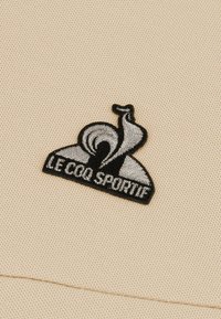 Tejido beige con un logotipo bordado en negro y gris que presenta un gallo estilizado y el texto "LE COQ SPORTIF" debajo.
