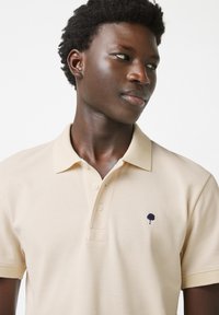 Polo beige con tessuto testurizzato, caratterizzato da un'apertura con tre bottoni e un piccolo emblema di un albero blu navy sul lato sinistro del petto.