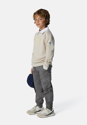 Felpa beige con la scritta "NORTH SAILS", pantaloni cargo grigi con tasche, camicia bianca con colletto visibile e sneakers color crema. Cappellino da baseball blu in mano.
