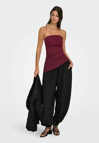 Burgundska majica brez naramnic z drapiranim dizajnom, v kombinaciji z ohlapnimi črnimi hlačami in črno jakno z čipkastim vzorcem, se dopolnjuje s črnimi sandali s trakovi.