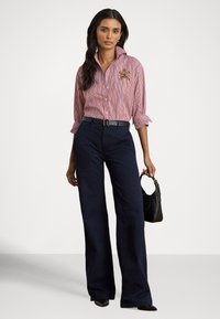 Sieviete, kas valkā sarkanu un zilu svītrainu pogājamu kreklu, tumši zilas platām kāju biksēm, melnu jostu, melnu rokassomu un melnas smailpurnu kurpes.