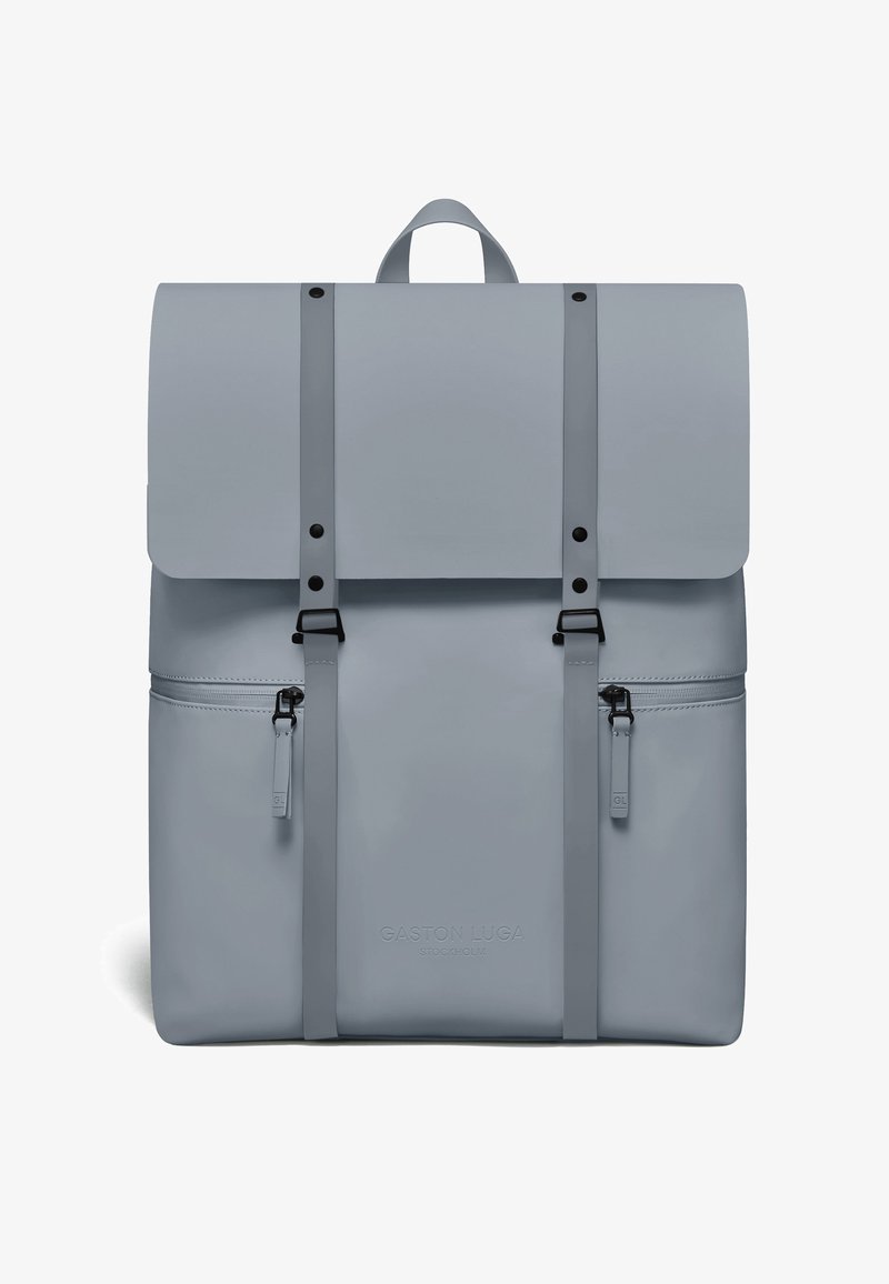 Sac à dos gris clair avec rabat supérieur, deux sangles verticales avec boucles, deux poches latérales zippées et une poignée de transport sur le dessus.