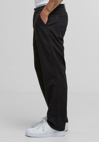 Person wearing black straight-leg pants, white sneakers, and a white shirt with a tattooed arm resting in pocket.