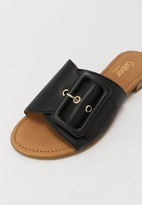 Musta nahkainen liukuva sandaali, jossa on leveä hihna, jossa on suorakulmainen solkiyksityiskohta, vaaleanruskea pohja ja minimaalinen ommel reunoilla.