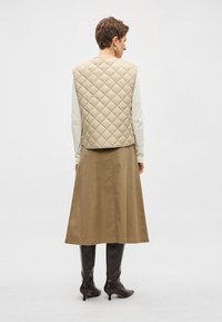 Colete matelassê bege sobre uma blusa de manga comprida creme, combinado com uma saia castanha até ao tornozelo e botas pretas até ao joelho. Design simples, sem padrões.