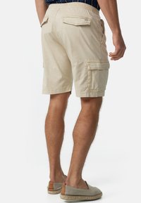 Man in beige cargoshorts en beige instap espadrilles, staande met één hand naast zijn lichaam tegen een effen achtergrond.
