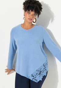 Top lungo a maniche con scollo a V di colore azzurro chiaro, con un accento di pizzo sull'orlo. Tessuto morbido con taglio asimmetrico.