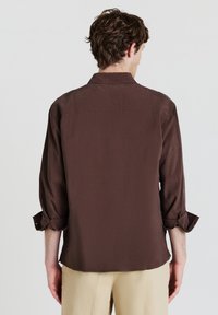 Uomo di spalle che indossa una camicia a maniche lunghe marrone scuro con i polsini risvoltati e pantaloni beige, davanti a uno sfondo chiaro e uniforme.