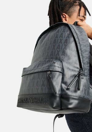 Zaino nero con logo in rilievo e materiale a fantasia, dotato di tasca frontale con zip e accenti testurizzati. Finitura liscia e design strutturato.