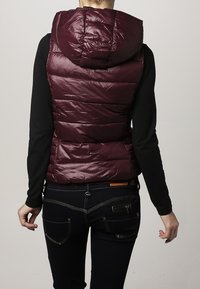 Bordeauxrote Steppweste mit Kapuze, mit gesteppter Struktur und glänzendem Finish. Über einem schwarzen Langarmshirt getragen und passt gut zu dunklen Jeans.