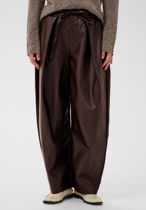 Weit geschnittene dunkelbraune Lederhose mit gebundenem Gürtel, kombiniert mit einem beigen Strickpullover und hellen flachen Schuhen.