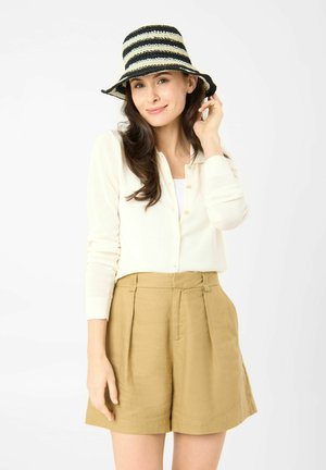 Femme portant un cardigan blanc, un short beige et un chapeau rayé noir et blanc, souriante et ajustant son chapeau d'une main.