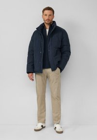 Dunkelblaue Daunenjacke mit hohem Kragen, Knopfverschluss und Fronttaschen, getragen über einem dunklen Pullover, khakifarbenen Hosen und weißen Sneakern.