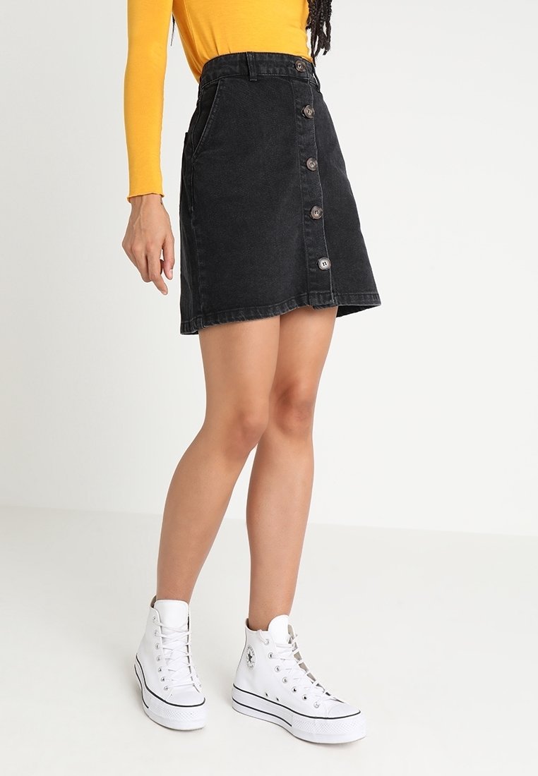 Jupe évasée en denim noir avec boutons sur le devant, taille haute, associée à des baskets montantes blanches. Texture lisse, design décontracté.