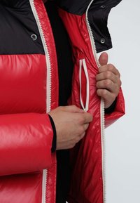 Red padded jacket with a shiny texture. Features a black collar, white zipper, and an internal pocket. Hands grasping the zipper.