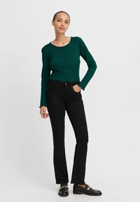 Grünes geripptes Langarmoberteil, kombiniert mit schwarzen geraden Jeans; Details sind der runde Ausschnitt und die fransigen Ärmelenden. Schwarze Loafer runden den Look ab.