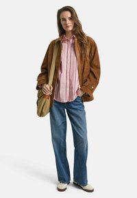 Brun mockaskinnjacka över en rosa randig skjorta med knappar, ihop med vida blå jeans och beige sneakers, samt en beige axelväska.