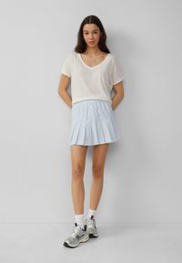 T-shirt blanc à manches courtes et col en V associé à une jupe plissée bleu clair avec des boutons sur le devant, portée avec des baskets grises et des chaussettes blanches.