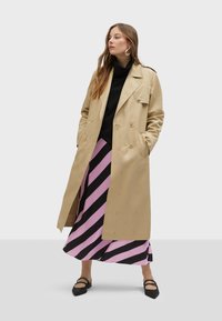 Beige trench coat with double-breasted design, epaulettes, and button detail, worn over a black turtleneck and pink-black striped skirt.