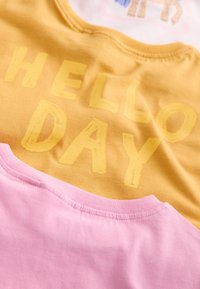 Rumeno oblačilo z besedilom "HELLO DAY" v zbledelo rumeni barvi, ki je na vrhu enostavnega rožnatega oblačila, pri čemer sta oba vidna šiva ovratnikov.