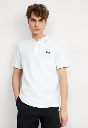Polo shirt bianca in cotone, con rifinitura nera al colletto e una piccola toppa nera con logo sul petto. Maniche corte, vestibilità classica.