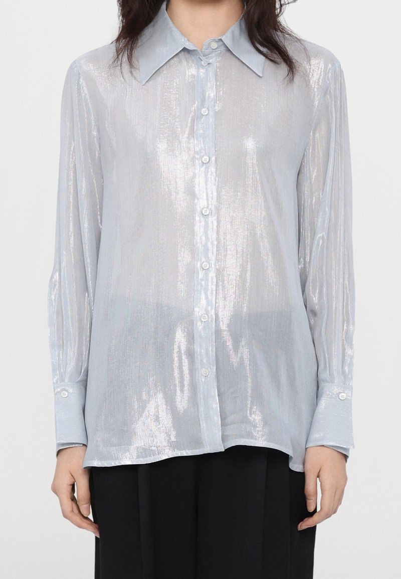 Femme portant une chemise brillante bleu clair à manches longues avec un col et des boutons, ainsi qu'un pantalon noir, debout devant un fond blanc.