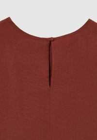Dettaglio ravvicinato di un colletto di un indumento color ruggine con un colletto rotondo e un piccolo bottone rivestito di tessuto sopra un'apertura a fessura.