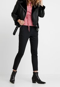 Giacca di pelle nera con colletto in pelliccia, blusa a righe rosse e bianche con cravatta, pantaloni neri aderenti e stivaletti neri con bordi sfrangiati.